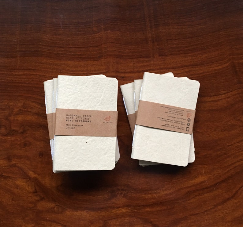 May include: Two stacks of handmade paper mini notebooks with brown paper covers. The cover of the stack on the left has the text "Handmade Paper Hand Stitched Mini Notebooks" and "Mini Notebook Unlined".