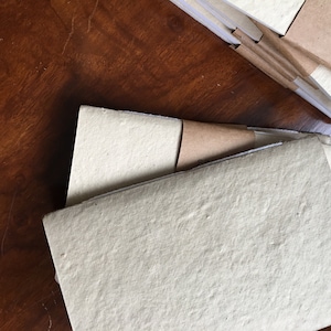 May include: A stack of blank, off-white paper notebooks with brown paper covers. The notebooks are bound with a simple stitch and have a rough, textured surface.