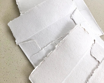 Puede incluir: Un montón de sobres de papel blanco hechos a mano con bordes rasgados. Los sobres están dispuestos en ángulo, mostrando su superficie texturizada y bordes irregulares únicos. Ideal para papelería o invitaciones.