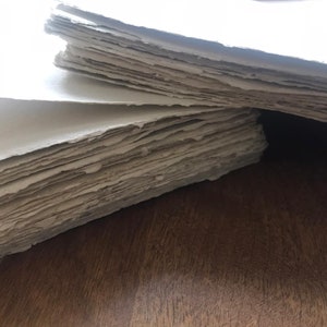 Puede incluir: Una pila de hojas de papel hechas a mano con una superficie rugosa y texturizada. El papel es de color beige claro y tiene un borde ligeramente irregular.