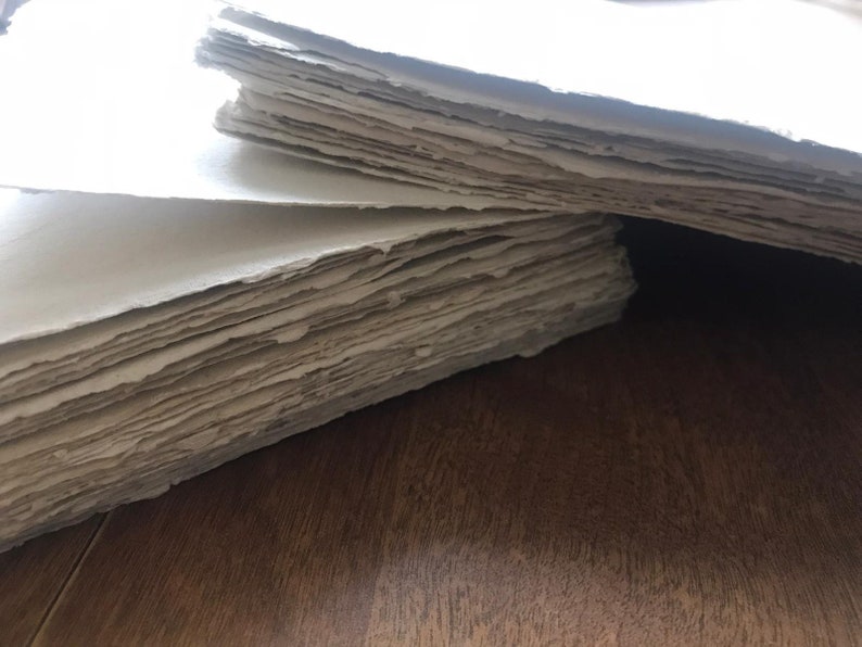 Puede incluir: Una pila de hojas de papel hechas a mano con una superficie rugosa y texturizada. El papel es de color beige claro y tiene un borde ligeramente irregular.