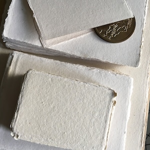 May include: A stack of handmade, off-white paper sheets with deckled edges. The paper has a textured surface and is perfect for crafting or writing.