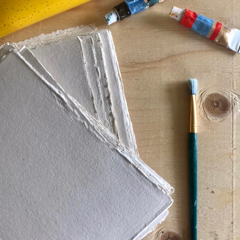 May include: A stack of handmade, off-white paper sheets with deckled edges. The paper is arranged on a light wood surface with a paintbrush and tubes of paint.