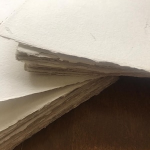 Puede incluir: Una pila de hojas de papel blanco hecho a mano con una superficie rugosa y texturizada. El papel est&aacute; dispuesto en una pila ligeramente irregular, con la hoja superior doblada.