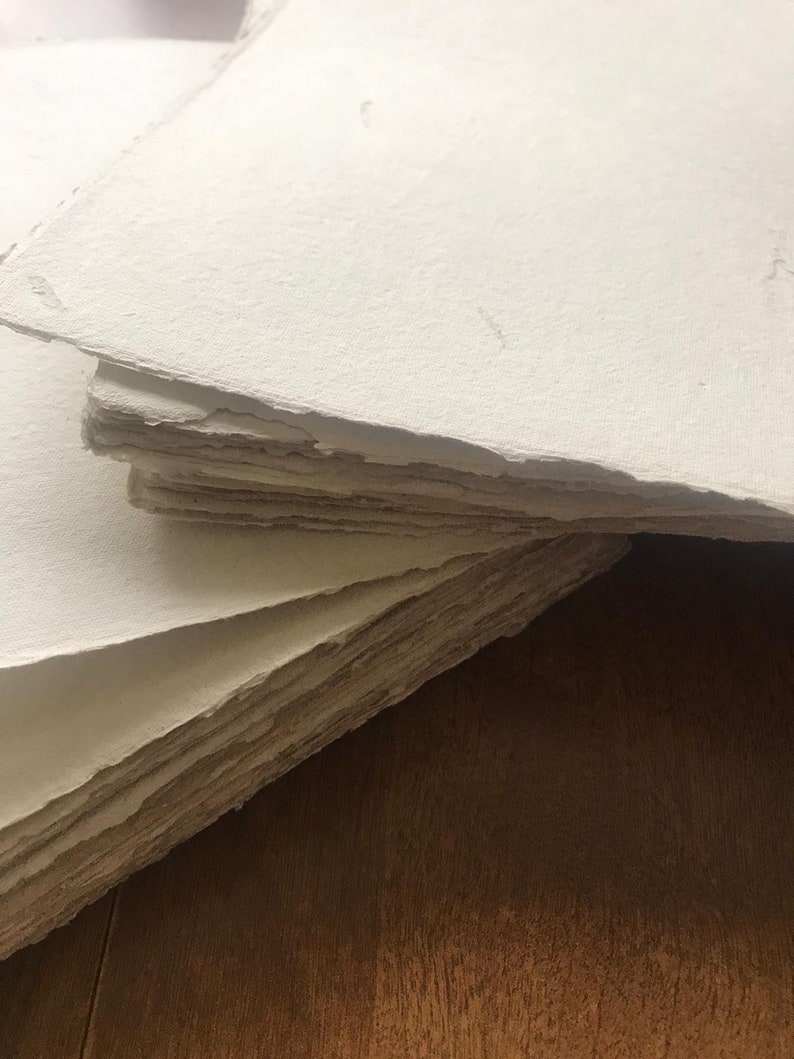 Puede incluir: Una pila de hojas de papel blanco hecho a mano con una superficie rugosa y texturizada. El papel est&aacute; dispuesto en una pila ligeramente irregular, con la hoja superior doblada.