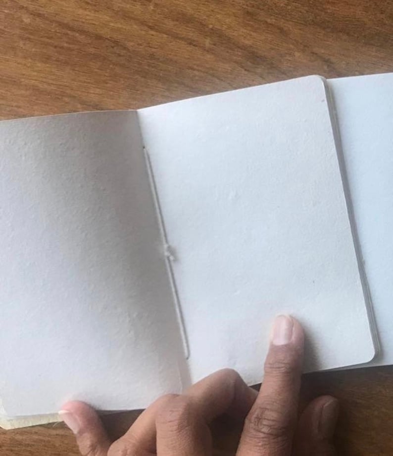 May include: A blank, white, paper notebook with a sewn binding. The notebook is open to a blank page.
