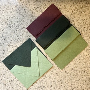 Op de afbeelding: Vier handgemaakte papieren enveloppen in verschillende kleuren: bordeauxrood, donkergroen, lichtgroen en een lichter groen met een gespikkelde textuur. De enveloppen zijn op elkaar gestapeld.