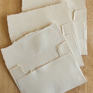 May include: Four white handmade paper envelopes with deckled edges. The envelopes are stacked on top of each other, with the largest envelope on the bottom and the smallest envelope on top.