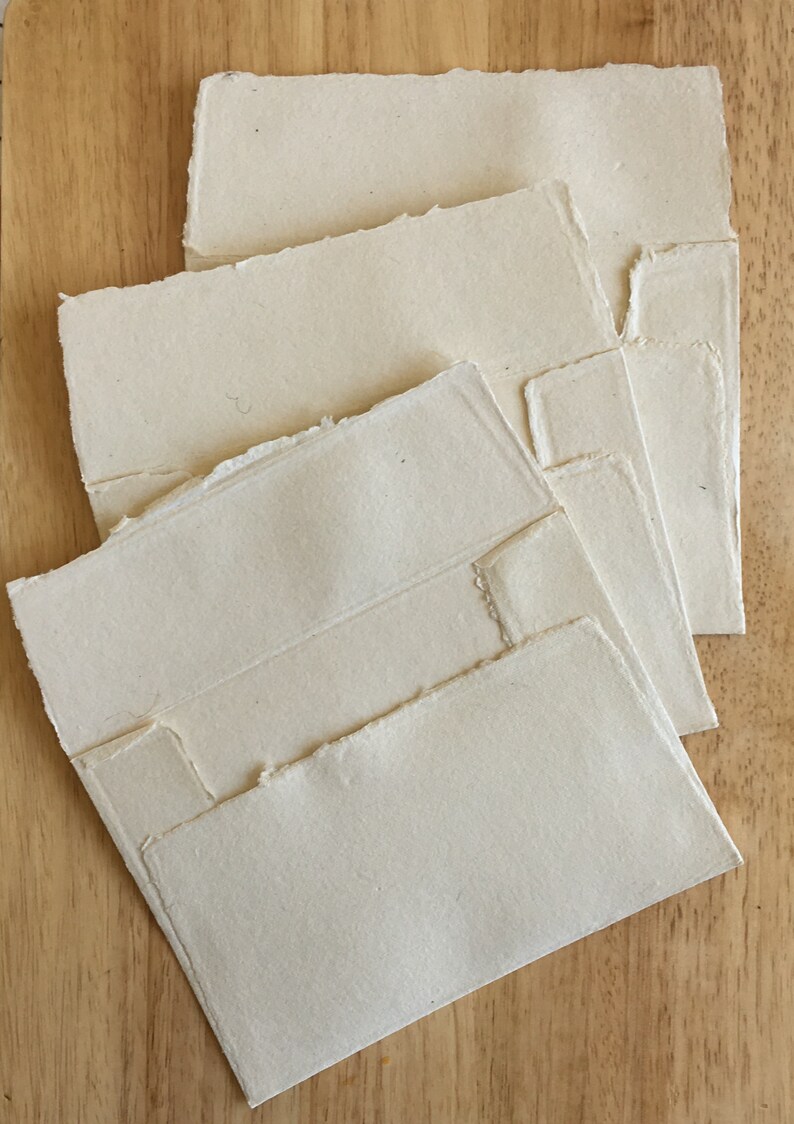 May include: Four white handmade paper envelopes with deckled edges. The envelopes are stacked on top of each other, with the largest envelope on the bottom and the smallest envelope on top.