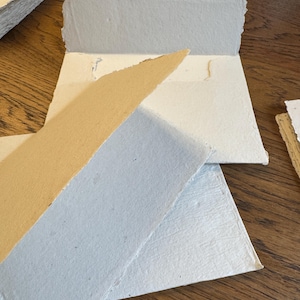 May include: A collection of handmade paper envelopes and sheets featuring deckled edges. The set includes various colors such as off-white, light blue, and tan. Each piece displays a textured, fibrous surface characteristic of artisanal paper production.