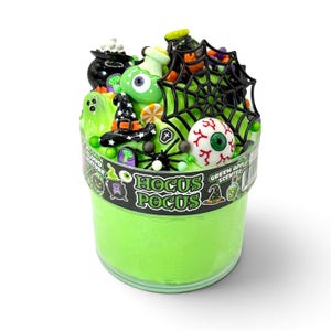 May include: A clear plastic container filled with bright green slime, topped with miniature Halloween-themed charms including a black spiderweb, a white eyeball, a black witch hat, and a ghost. The label reads Hocus Pocus and Green Apple Scented.