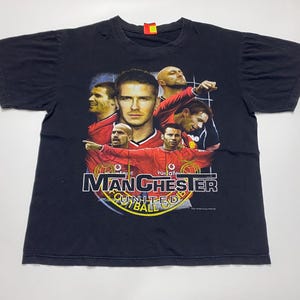 Puede incluir: Camiseta negra con un gráfico de jugadores de fútbol del Manchester United en rojo. La camiseta tiene las palabras "Manchester United Football" en blanco y amarillo. La camiseta está hecha de un material suave.