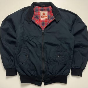 Può includere: Una giacca blu navy con cerniera e fodera a quadri rossi, verdi e blu. La giacca presenta un colletto, maniche lunghe con polsini a coste, un orlo a coste e due tasche frontali con chiusura a bottone. L'etichetta interna riporta Baracuta Made in England.