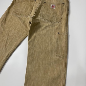 May include: Tan work pants featuring a back cinch strap, two large rear patch pockets, and a side utility pocket. A white and red Super SB brand patch is sewn onto the right rear pocket, and a rectangular brand label is located at the waist.
