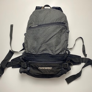 May include: Black and gray backpack with a grid pattern. The backpack has multiple compartments, adjustable straps, and a waist belt. The word "HYSTERIC" is printed on the front. The backpack is designed for outdoor activities.