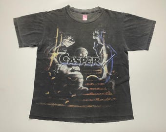 May include: Dark gray t-shirt featuring a graphic of Casper the Friendly Ghost and his ghostly friends. The word "Casper" is printed in a blue and white font. The shirt has a vintage, worn look.