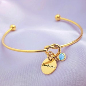 Puede incluir: Pulsera de brazalete de oro con un dije de nudo y un pequeño dije redondo con el nombre "Isabella" grabado. Una piedra preciosa azul claro está unida al dije de nudo.