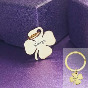 Puede incluir: Un dije de trébol de cuatro hojas de color dorado con el nombre "Robyn" grabado. El dije está unido a un llavero dorado. El trébol tiene forma de trébol de cuatro hojas. El fondo es una superficie morada texturizada.