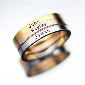 May include: Three stacked rings with the names "John", "Hayley", and "James" engraved on them. The rings are made of different metals: gold, rose gold, and silver.