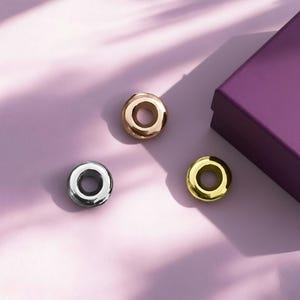 Può includere: Tre perline circolari metalliche, oro, argento e oro rosa, sono disposte su una superficie lilla chiaro. Sullo sfondo è presente una scatola viola parzialmente visibile. Le perline hanno una finitura liscia e lucida e un foro al centro.