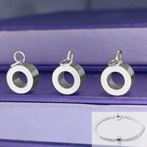 May include: Three silver-colored, circular charm beads with a small loop at the top. The beads have a thick, cylindrical shape with a large hole in the center. A silver bracelet with a clasp and charm is shown at the bottom.