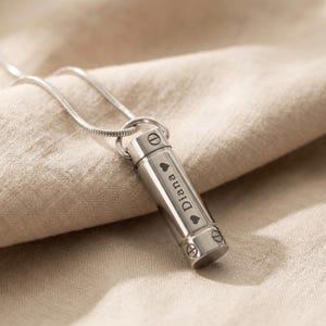 Op de afbeelding: Een zilverkleurige, cilindervormige hangerketting. De hanger is gegraveerd met de naam "Diana" en hartsymbolen. De ketting hangt aan een dunne, zilveren ketting. De achtergrond is een neutraal gekleurde stof.