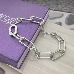 Puede incluir: Una pulsera de eslabones plateados con eslabones ovalados alargados. La pulsera está sobre una caja morada. La pulsera es de metal brillante y tiene un diseño sencillo y moderno. La palabra "FEVER" es visible en la caja.