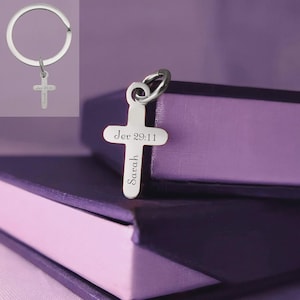 以下が含まれることがあります： 「Jer 29:11」と「Sarah」の文字が刻まれた、銀色の十字架型のキーホルダー。キーホルダーは銀色のリングに付いています。背景は紫色のグラデーションです。