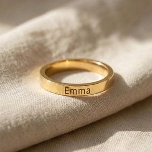 May include: A polished gold-tone metal band ring featuring a flat outer surface. The name Emma is engraved in a clean, sans-serif black font on the front of the ring. The band has a smooth, reflective finish and a consistent width throughout its circular shape.