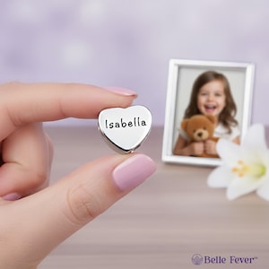Op de afbeelding: Een kleine, hartvormige, zilveren bedel met de naam "Isabella" gegraveerd. De bedel wordt vastgehouden voor een ingelijste foto van een lachend kind met een teddybeer. Een witte bloem staat op de achtergrond.