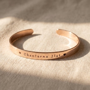 May include: A rose gold cuff bracelet with the engraved words "Chantarna 21st" and heart symbols. The open bangle is made of a shiny, metallic material. The bracelet is displayed on a neutral-toned fabric.