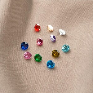 May include: A collection of eleven small, round, faceted gemstones in a variety of vibrant colours including red, blue, green, pink, purple, yellow, and clear. Each stone features a polished, multi-faceted surface that reflects light.