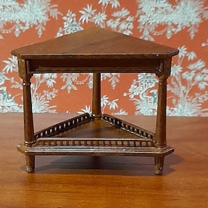 May include: A miniature, wooden, triangular table in a dark brown hue. It has three legs and a decorative shelf. The background showcases patterned wallpaper in orange and white tones.