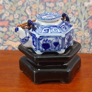 May include: A small blue and white porcelain teapot with a lid and a gold handle, sitting on a black hexagonal stand.