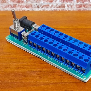 May include: A green circuit board with two rows of blue screw terminals labelled with numbers 1 through 12. A toggle switch and a black power jack are also on the board.