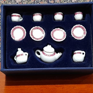 May include: A miniature tea set in a blue box. The set includes a teapot, creamer, sugar bowl, four cups, and four saucers. The porcelain is white with pink and gold trim.