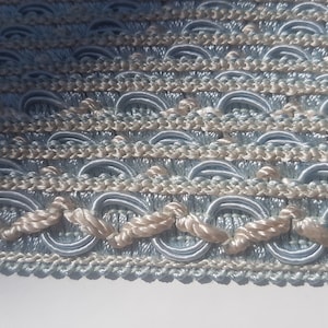 May include: Close-up of a light blue and beige braided trim with a decorative pattern. The trim is made of woven fabric and has a rope-like texture.