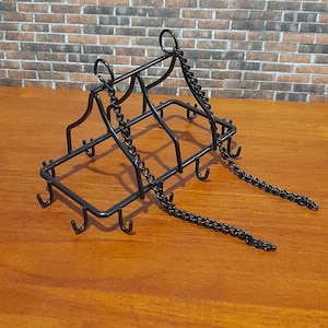May include: A black metal pot rack with hooks and chain details. The rack is designed to hang pots and pans. The rack is on a wooden surface with a brick wall background.