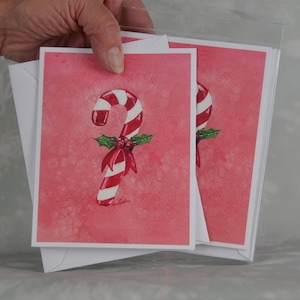 Può includere: Un set di biglietti di auguri con un dipinto ad acquerello di un bastoncino di zucchero a strisce rosse e bianche, adornato con un fiocco rosso e agrifoglio. I biglietti hanno uno sfondo rosa e sono tenuti in una custodia di plastica trasparente con buste bianche.