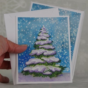 Può includere: Biglietti di Natale ad acquerello con un albero di Natale innevato. L'albero è dipinto in tonalità di verde e viola, con neve bianca. Lo sfondo è azzurro con fiocchi di neve bianchi. I biglietti sono abbinati a buste bianche.