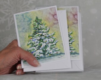 May include: Watercolour Christmas cards depicting a snow-covered fir tree with colourful baubles. The cards have a white border and are held in a clear plastic sleeve. The artwork has a soft, painterly style.