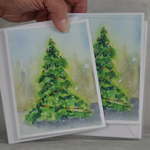 Può includere: Due biglietti di auguri natalizi con un dipinto ad acquerello di un albero di Natale decorato. L'albero è verde con luci colorate su uno sfondo innevato azzurro e verde. I biglietti sono in buste bianche.