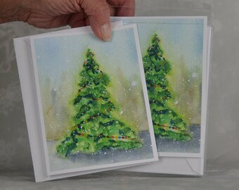 May include: Two Christmas cards with a watercolour painting of a decorated evergreen tree. The tree is green with colourful lights against a snowy, light blue and green background. The cards are in white envelopes.