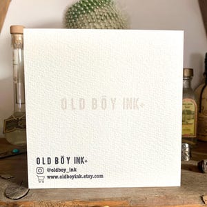 May include: White card with the text "OLD BOY INK" in a light pink font. The text "OLD BOY INK" is repeated in a smaller font at the bottom of the card, along with the social media handle "@oldboy_ink" and the website "www.oldboyink.etsy.com".
