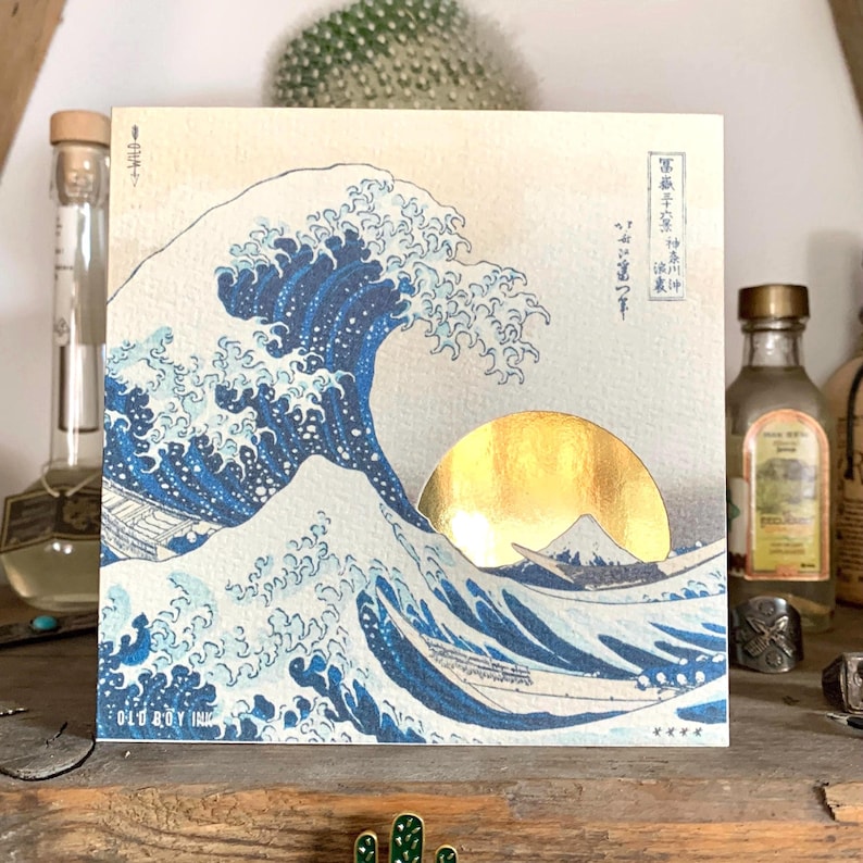May include: A vintage-style print of the Great Wave off Kanagawa by Katsushika Hokusai. The wave is depicted in shades of blue and white, with a golden sun rising behind it. Mount Fuji is visible in the background. The print is framed with a white border and has the text "Old Boy Ink" at the bottom.