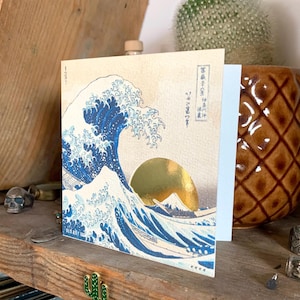 May include: A greeting card featuring a blue and white print of a large wave with a gold sun and mountain in the background. The Japanese text on the card reads "神奈川沖浪裏" which translates to "The Great Wave off Kanagawa".