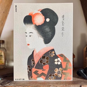 May include: A print of a geisha in profile, featuring a traditional hairstyle with a coral-colored flower. The geisha wears a black kimono with floral patterns and a coral and gold design. The print has Japanese text and the text "OLD BOY INK-".