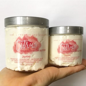 May include: Two glass jars of whipped body butter labeled "Citrus Whipped Body Butter". The jars have silver lids and are filled with a white, creamy product. The labels are pink and white with a watercolor-style design. The ingredients are listed on the labels: Shea butter, cocoa butter, coconut oil, olive oil, grapefruit essential oil, sweet orange essential oil, lemon essential oil. Organic ingredients.