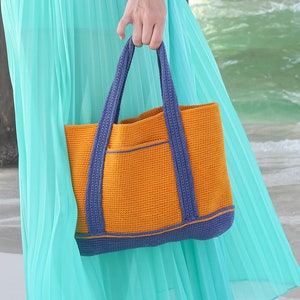 May include: A woman wearing a white top and a turquoise maxi skirt is standing on a sandy beach. She is holding a bright orange tote bag with blue handles.