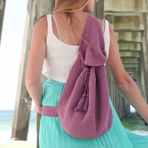 Może przedstawiać: Kobieta w białym topie na ramiączkach i długiej, turkusowej spódnicy stoi pod pomostem. Ma na sobie plecioną, fioletową torbę z sznurkiem.